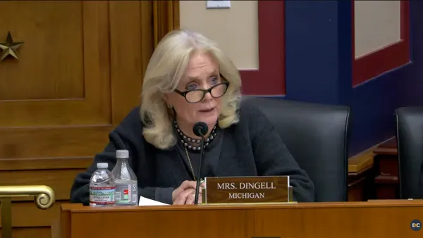 A woman wearing glasses sits at a dais with a sign reading, "Mrs. Dingell, Michigan."