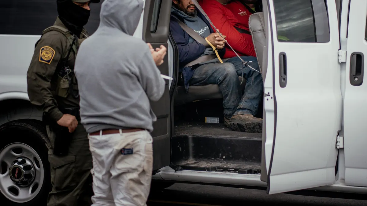Detained individuals sit in a vehicle while an immigration official stands nearby.