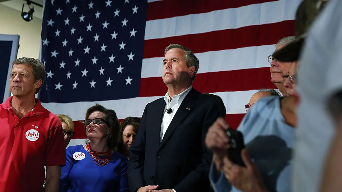 Jeb Bush is standing the center of a handful of people who have Jeb! stickers on their clothes. A large American flag is on the wall behind the people.