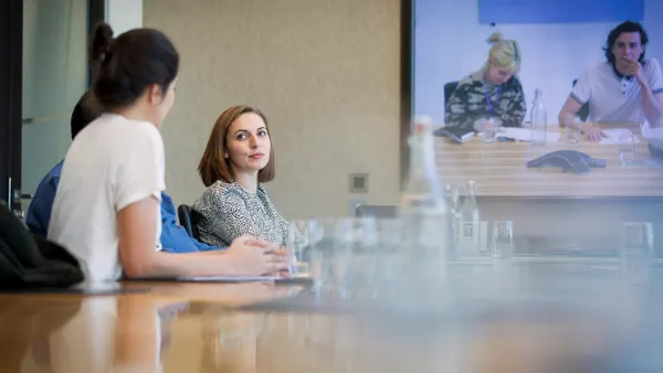 A group of professionals in a conference room having a meeting, with a video conference screen showing two additional participants on a call.