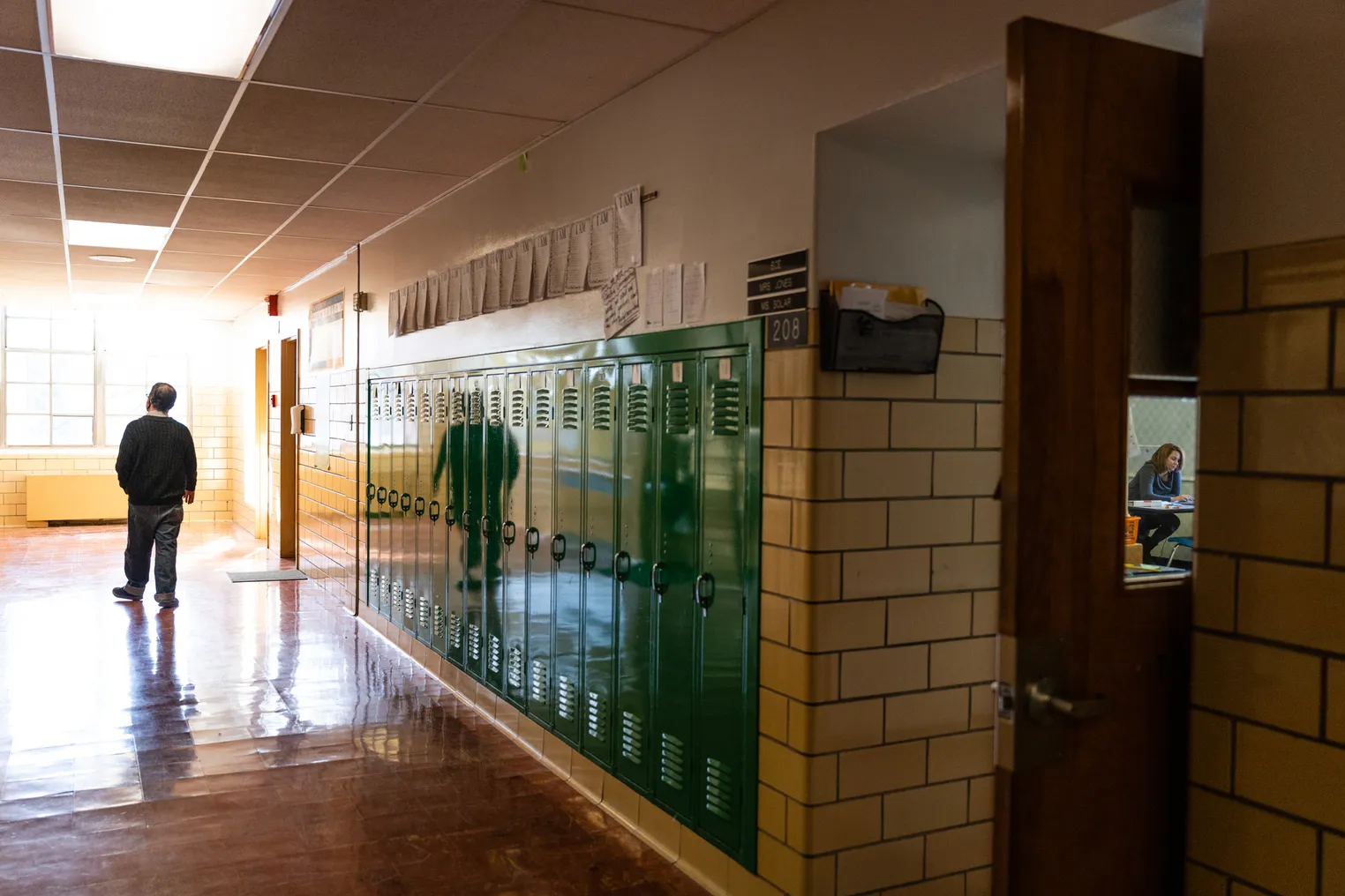 A person walks down a school hallway lined with lockers as another person sitting in a classroom can be seen through a window.