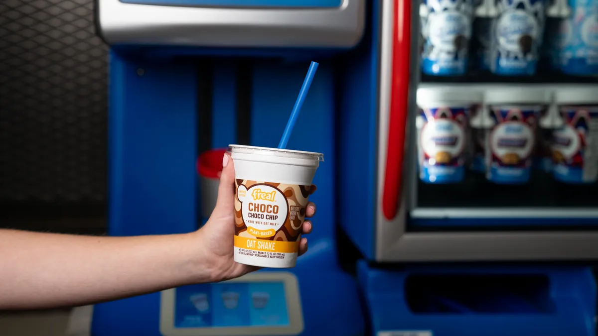 A photo of a hand holding a plastic cup with a mixing machine in the background. The cup says "f'real choco choco chip oat shake."