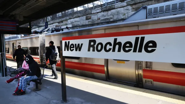 Several people on a train station platform with a commuter train and a sign reading "New Rochelle."