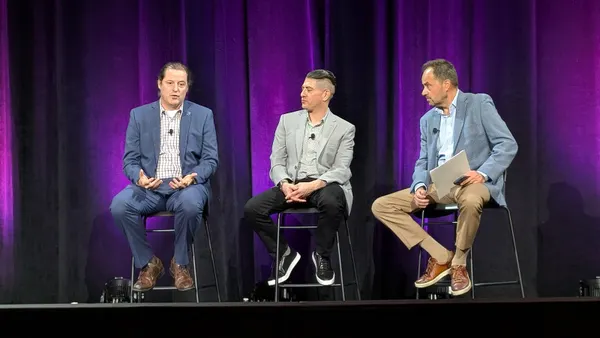 Three men sit on chairs on a stage. One man gestures with his hands while the other two look at him.