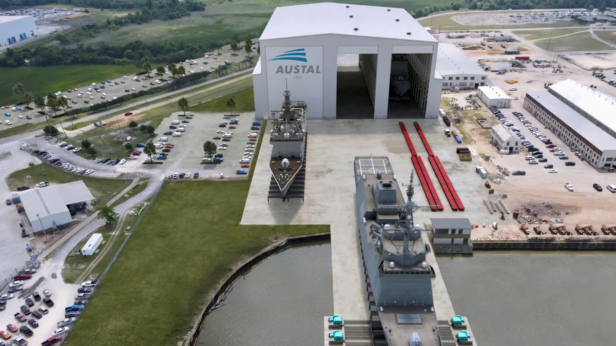 A white building with the gray and blue Austal logo on it, and a long gray ship in front of the building.