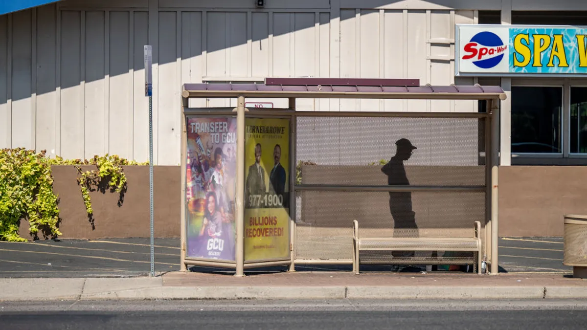 A silhouette of a person wearing a hat under a bus stop on the side of the road. Behind the bus stop is a building.