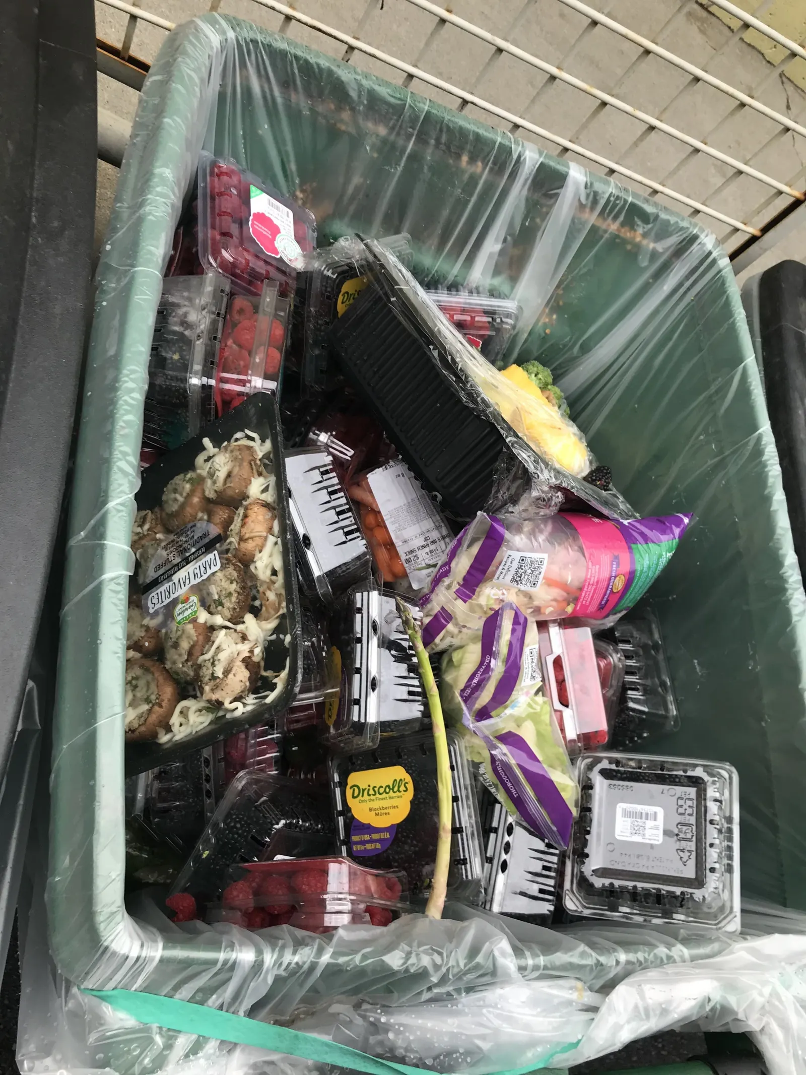 A waste bin with a liner is filled with packaged food, including berries, mushrooms and bagged salad kits.