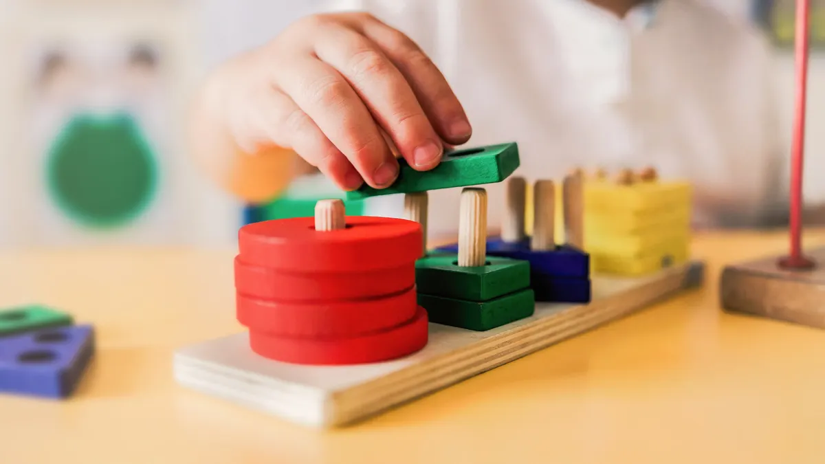 A child's hand is seen moving a colorful block to a peg wooden board.