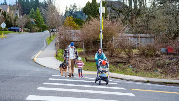 Two adults, a young child and baby in a stroller, accompanied by a dog, cross the street amond trees and homes in the background.