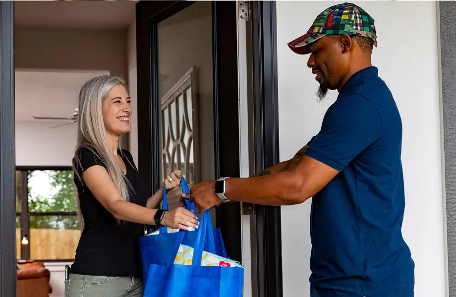 Person hands two blue bags to person at the front door of a residence.