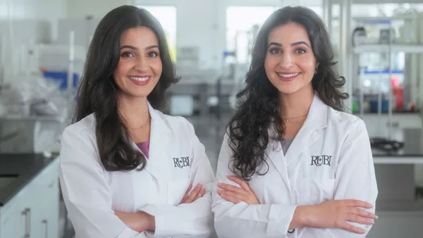 Two women in white lab coats that read "Rubi" stand with crossed arms and smile at the camera.