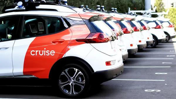 A fleet of Cruise ride-hailing vehicles parked in a row outdoors on a sunny day with buildings and greenery in the background.