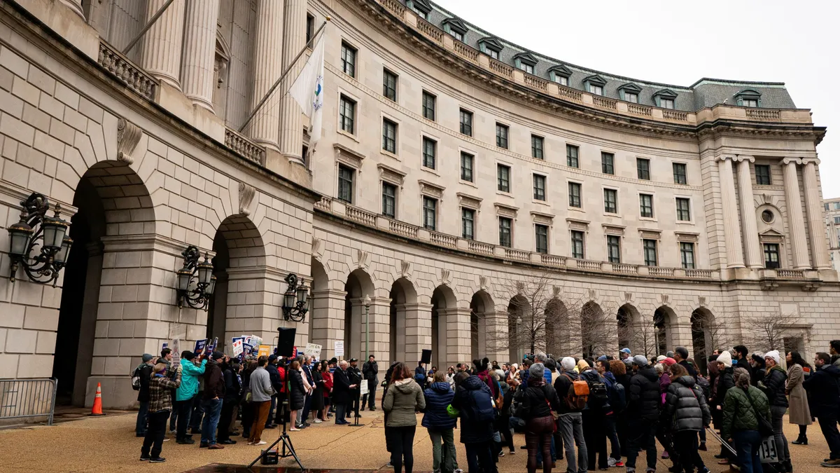 A group of protesters in winter clothing stand near a speaker system outside a large neoclassical building.