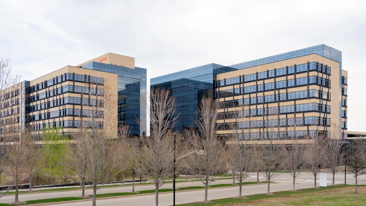 A modern office building with a double glass facade is lined with bare trees in the foreground. A sign reads "Optum" on the building's left side.