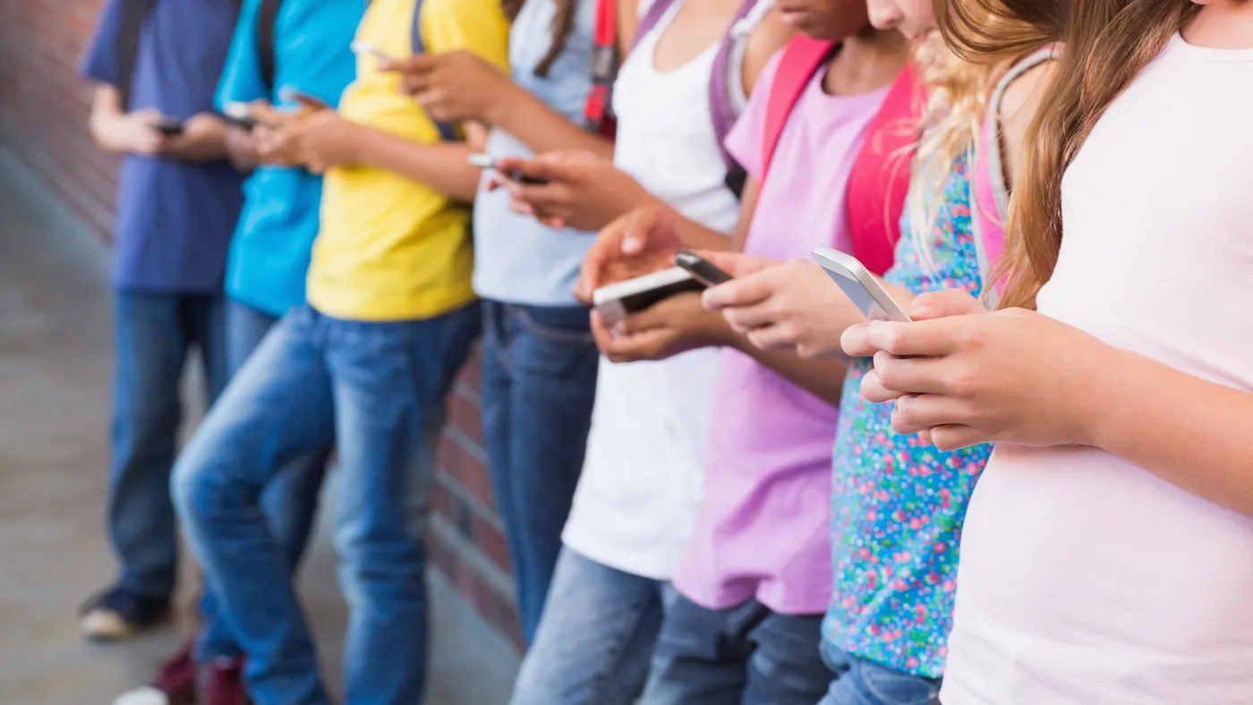 A row of students standing and leaning against a brick wall. In their hands are cellphones.