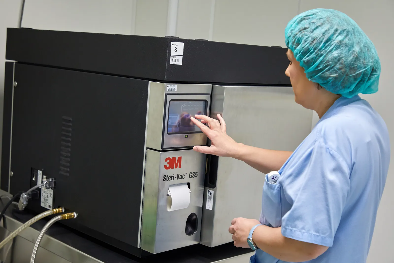 A person wearing scrubs and a hairnet taps a screen on a square machine that says "3M Steri-Vac GS5."