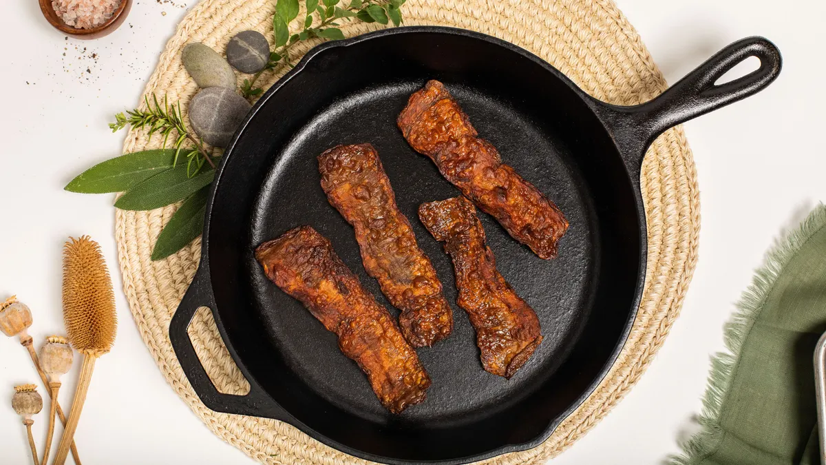 A cast iron skillet with four cooked slices of MyBacon sits on a beige round woven placemat with herbs and stones at the upper left, as well as a small dish of pink salt.