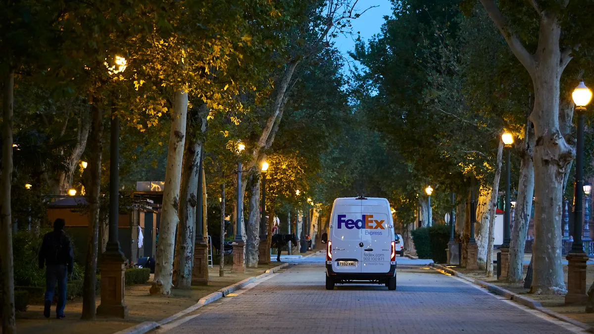 A FedEx Express delivery vehicle with European plates driving down a tree-lined street.