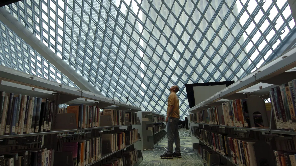 A person stands in between bookshelves looking up at a transparent ceiling, behind which are tall city buildings.