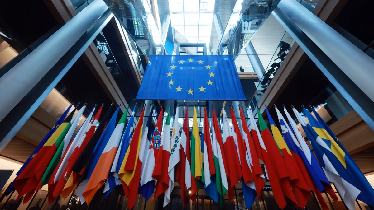 A group of flags on flag poles stand underneath one large blue flag, which features a circle of gold stars in the middle.