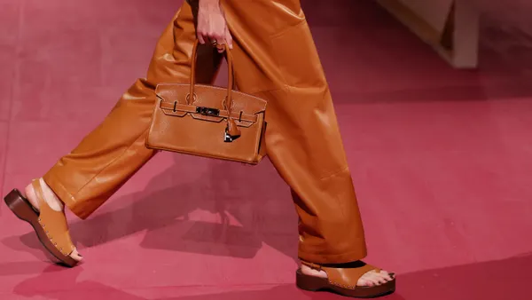 A sideview of a model in orange leather pants carrying an orange leather Hermès bag on a runway.
