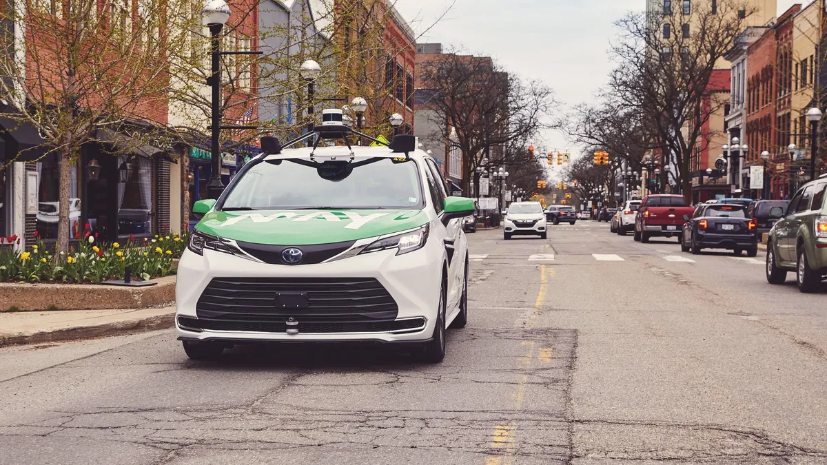 A frontal view of a white vehicle with a green hood, outfitted with rooftop cameras and sensors, driving along a busy street with brick buildings.