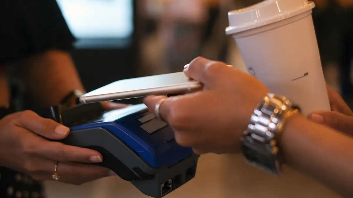 Close-up shot of unrecognizable female hands holding her smartphone above card reader while paying contactless for her takeaway coffee held in other hand