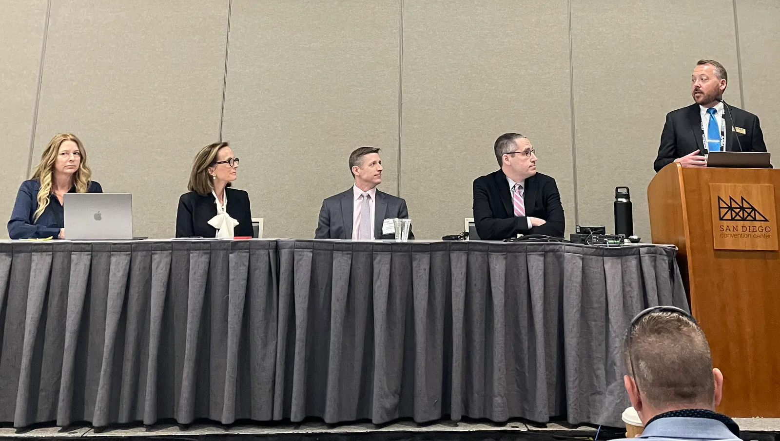 A group of five man and woman school administrators are seated at a table on a dais in a convention center meeting hall.