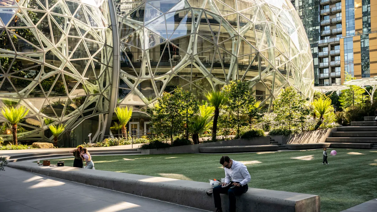 A handful of people sit and walk on a small green area next to large glass spherical buildings on a sunny day.