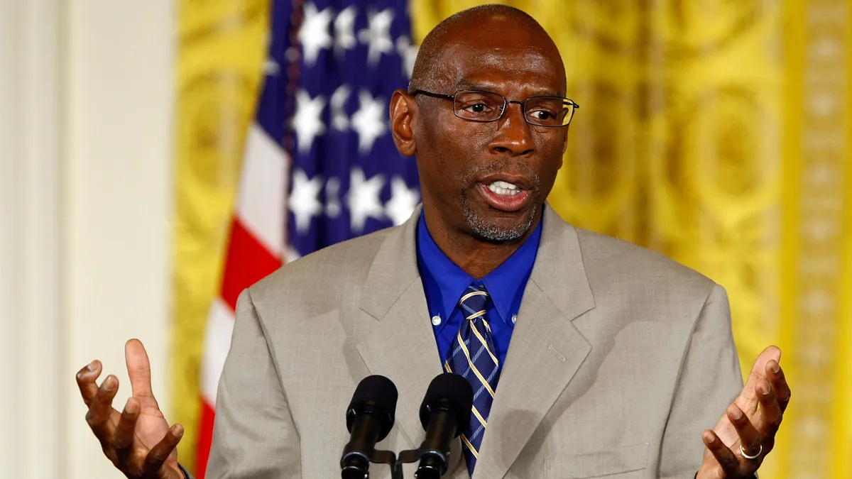 A Black professional man speaks at a podium during a White House event. A U.S. flag is visible but out of focus in the background.