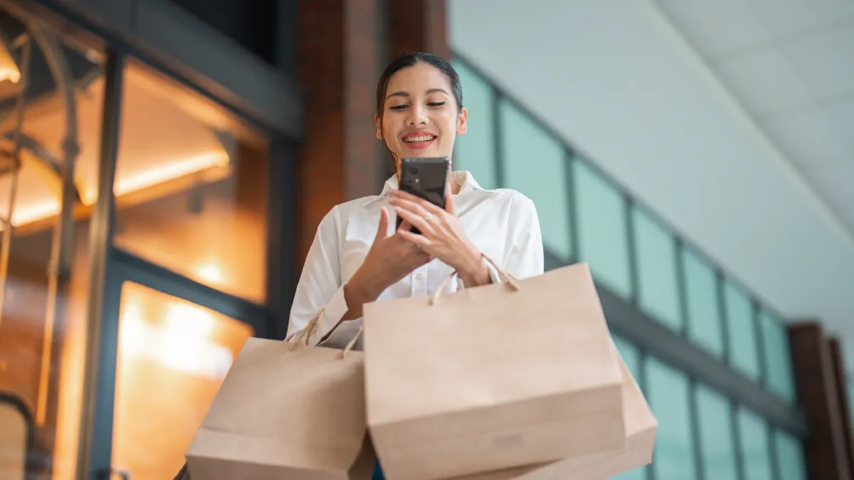 Woman online shopping through cellphone while at the mall, holding shopping bags