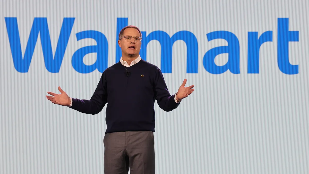 A man speaks while standing in front of a screen displaying "Walmart" in blue letters.