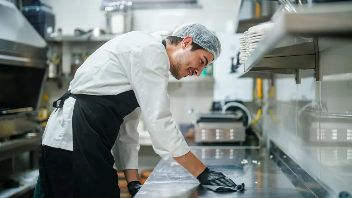 A photograph of a person wearing a white shirt and black pants and black apron. The person is wiping down a kitchen surface.
