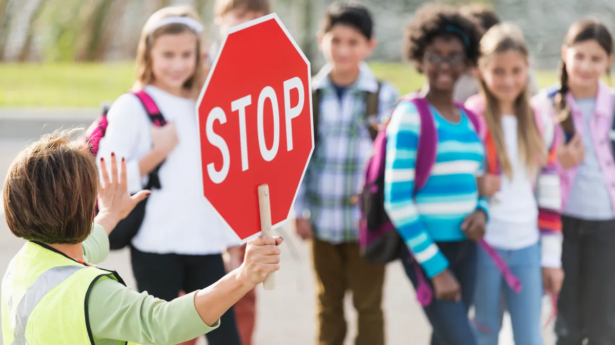 Students wait to walk across the street as a crossing guard tells them to wait while holding a stop sign.