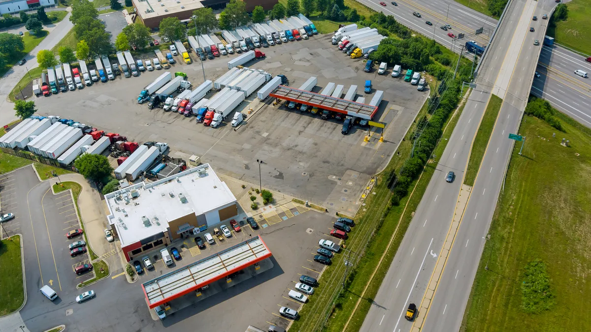 An aerial view shows dozens of trucks at a rest top next to a four-lane highway.