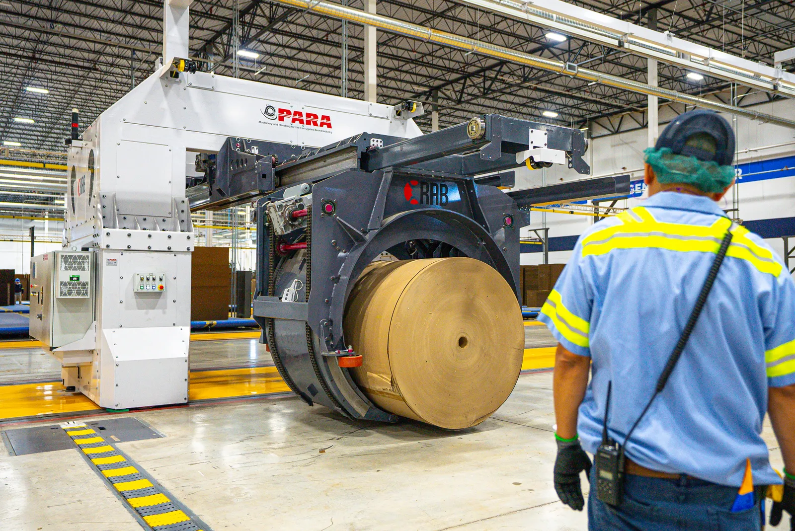A person wearing safety gear watches a large piece of equipment grab a paper roll inside an industrial facility.