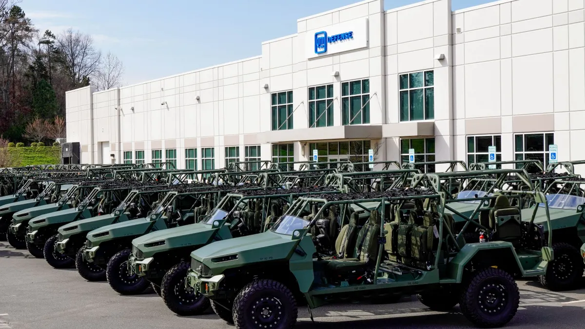 Two rows of green infantry squad vehicles parked in front of a white building with the blue GM Defense logo on it.