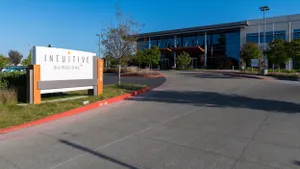 A modern office building with large reflective glass windows is shown behind a wide driveway and landscaped entrance. In the foreground, a white and orange monument sign reads "Intuitive Surgical" with the number "1020" vertically on the side.