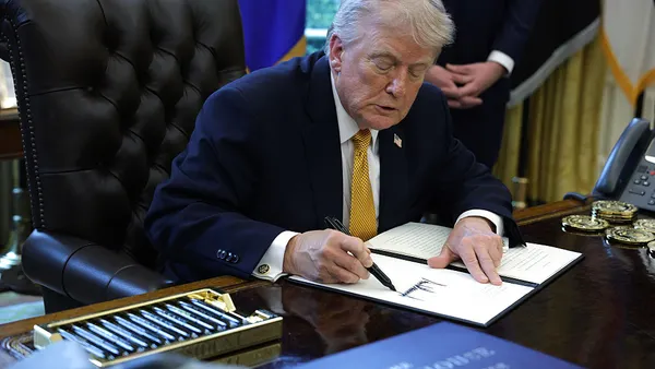 Wearing a suit and gold tie, President Donald Trump leans forward over a desk with a pen and signs an executive order.