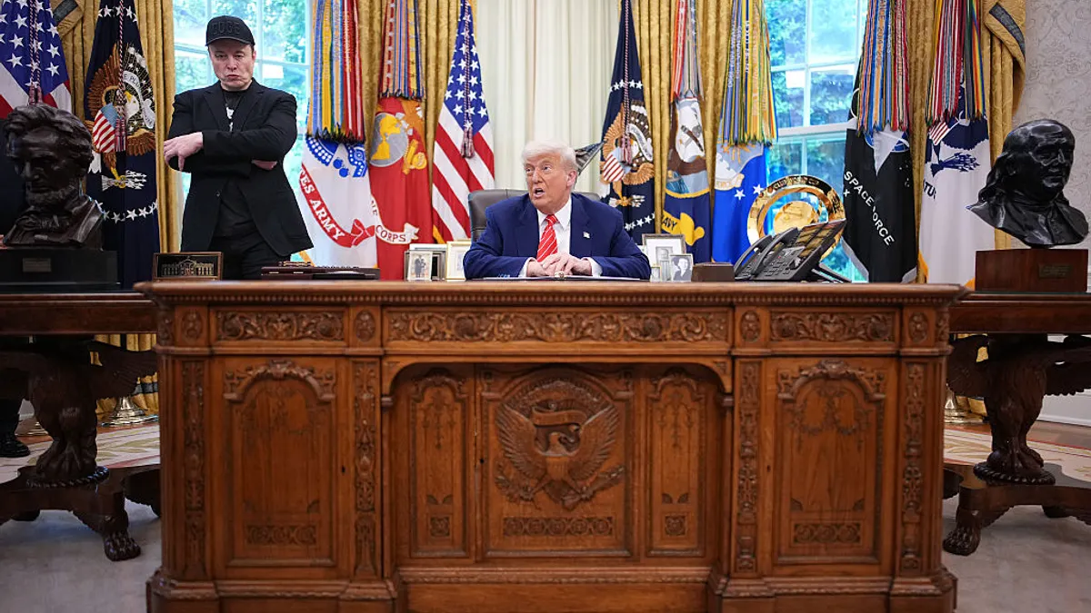 Two people are in the Oval Office at the White House. One person is standing and the other is sitting at a desk.