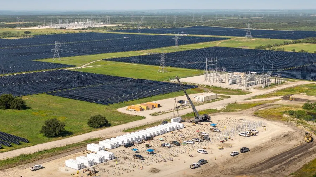 An aerial view of solar panels in a field with transmission lines and a crane installing large containers of battery storage in a dirt lot.