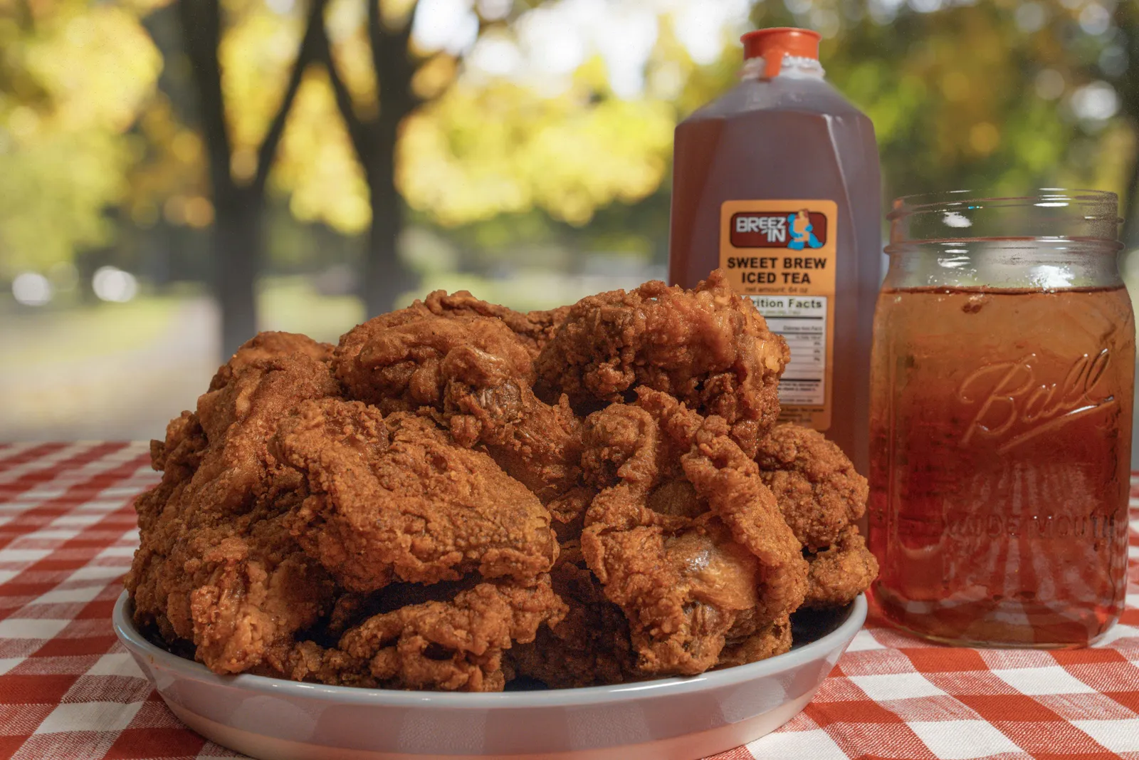 A photo of a place of fried chicken on a table outdoors with a red checkered tablecloth under it and a glass and jug of tea behind it.