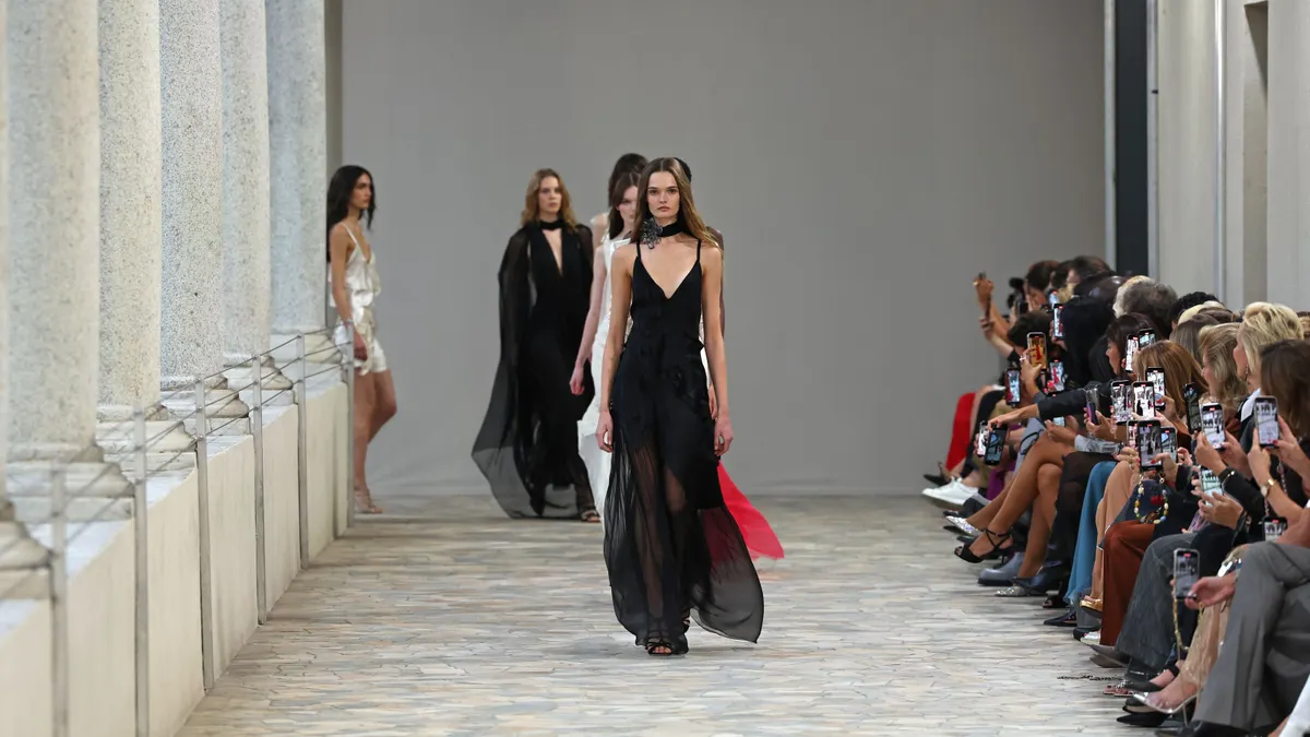 Models in flowing, long dresses walk down a runway as a seated crowd applauds from the side.