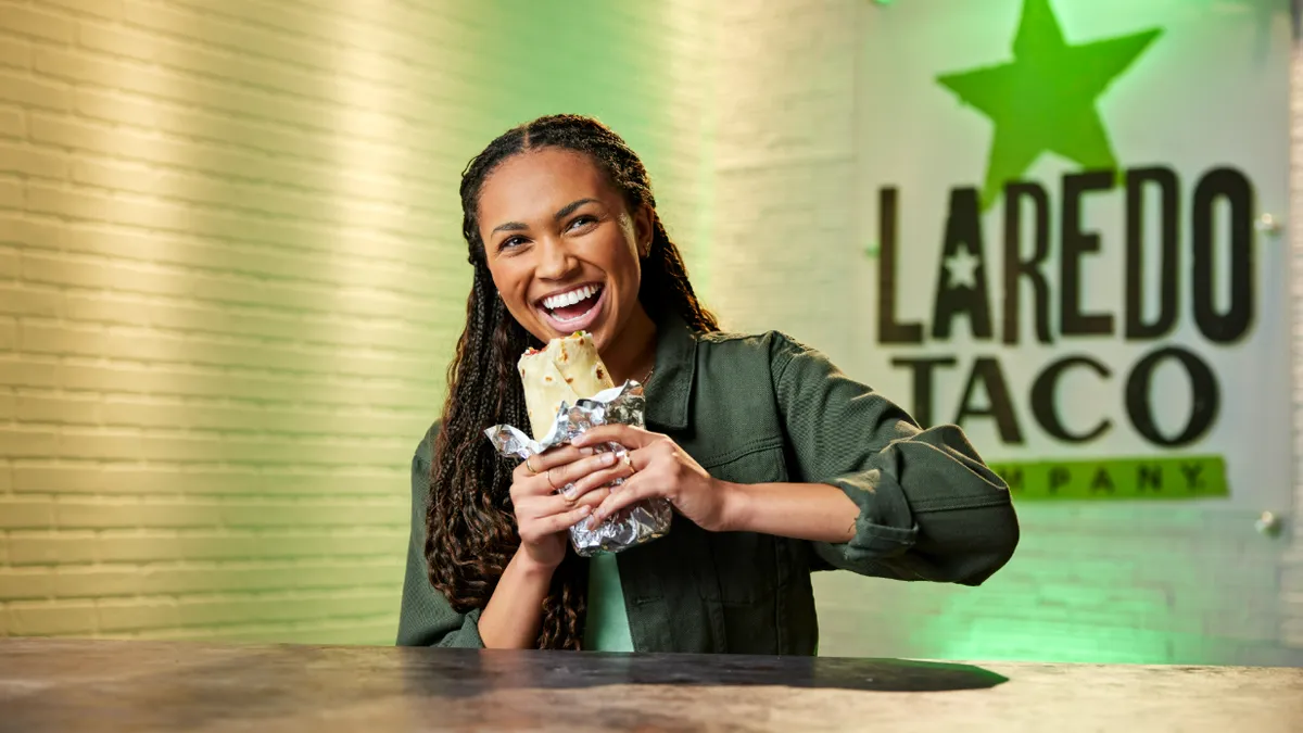 A photo of a smiling person sitting at a table, about to bite into a burrito. A sign on the wall behind them says Laredo Taco.