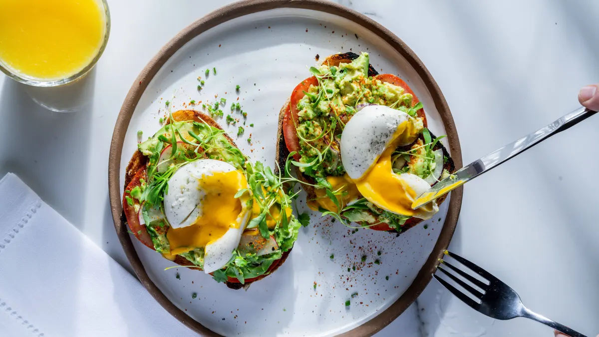 Overhead view of two slices of avocado toast, each topped with a cut Yo Egg, sitting on a white plate with a tan border. A person is holding a knife and fork to the right.
