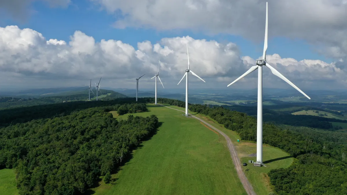 Wind turbines stand against a backdrop of green fields and mountains.