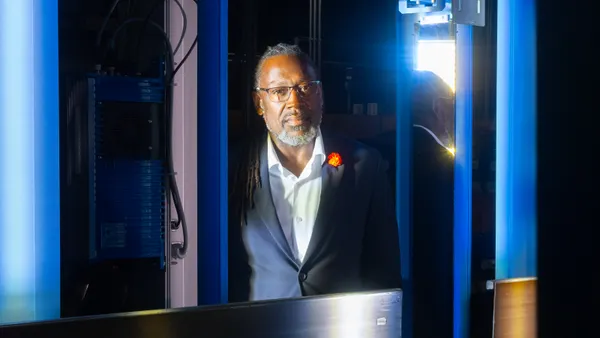 A person with glasses wearing a dark suit jacket stands for a portrait, framed between the blue lights of industrial warehouse machinery.
