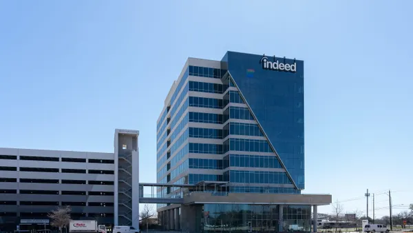 A tall glass office tower labeled “indeed” next to a multi-level parking garage and stair tower, with blue sky in the background.