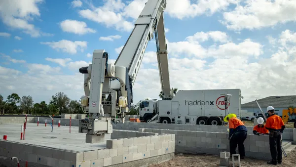 A robot arm moves cinderblocks from the back of a truck with the assistance of construction workers.