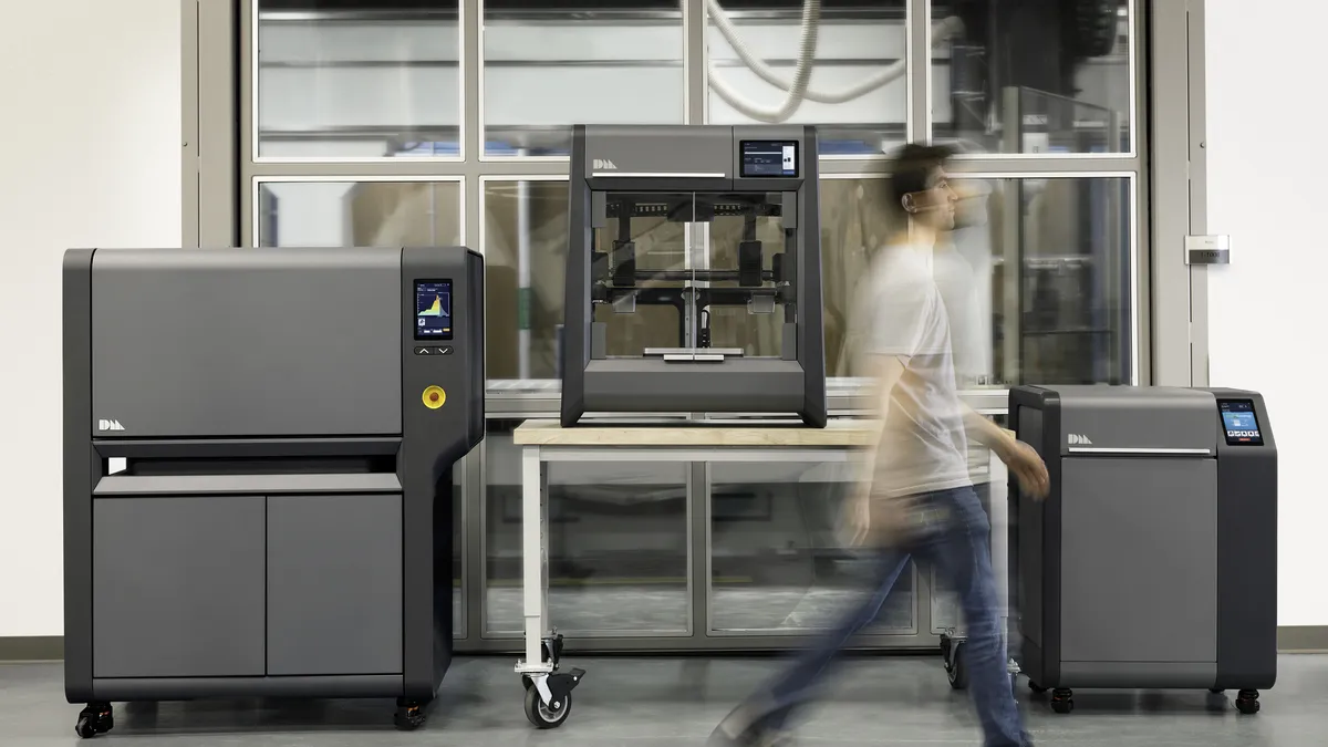 A dark gray additive manufacturing system with reflective dark glass windows behind it and a blurry person walking in front of it.